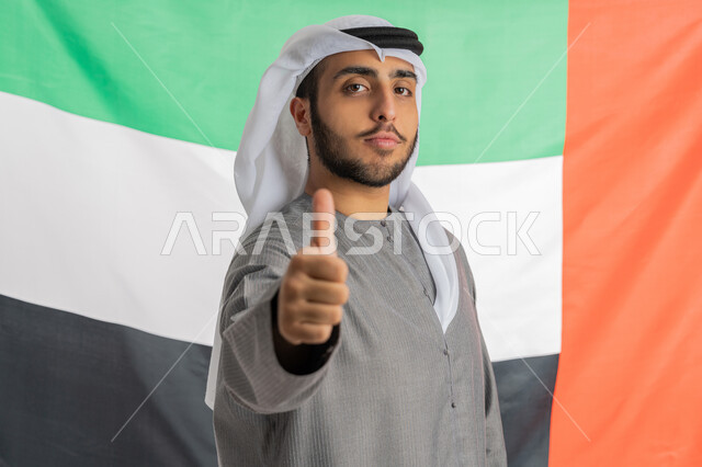 ذكرى يوم الاتحاد الوطني الإماراتي 2 ديسمبر، صورة لشاب إماراتي عربي خليجي يرتدي الزي التقليدي ينظر إلى الكاميرا ويرفع يده بايماءات الإعجاب والفخر بالانتماء للوطن وخلفه العلم الإماراتي، الاحتفال بيوم العلم الإماراتي 3 نوفمبر، الأعياد والمناسبات الوطنية