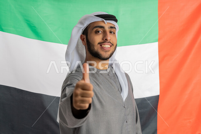 يوم الاتحاد والاستقلال الإماراتي 2 ديسمبر، صورة لشاب إماراتي عربي خليجي مبتسم يرتدي الزي التقليدي ينظر إلى الكاميرا ويرفع يده بايماءات الفخر والإعجاب بالانتماء للوطن وخلفه العلم الإماراتي، الاحتفال بيوم العلم الإماراتي 3 نوفمبر، الأعياد والمناسبات الوطنية