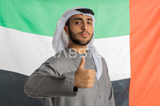 ذكرى يوم الاتحاد والاستقلال الإماراتي 2 ديسمبر، صورة لشاب إماراتي عربي خليجي يرتدي الزي التقليدي ينظر إلى الكاميرا ويرفع يده بايماءات الإعجاب والافتخار بالانتماء للوطن وخلفه العلم الإماراتي، الاحتفال بيوم العلم الإماراتي 3 نوفمبر، الأعياد والمناسبات الوطنية