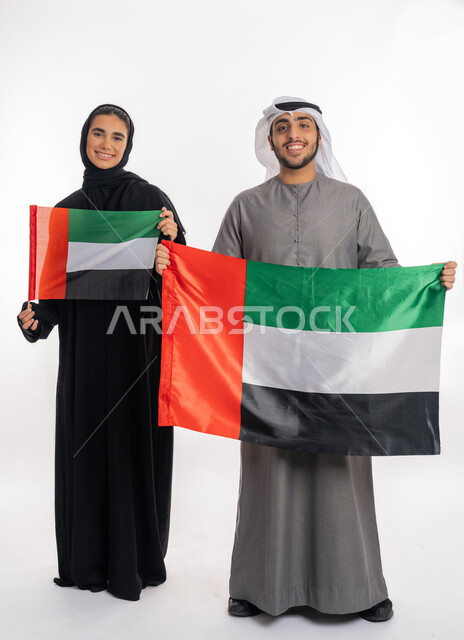 الاحتفال باليوم الوطني الإماراتي٢ ديسمبر، بورتريه لشاب وفتاة عربيان خليجيان إماراتيان مبتسمان يرتديان الزي التقليدي ويحمل كل منهما علم الإمارات بحجمين مختلفين، ذكرى يوم العلم الإماراتي ٣ نوفمبر، الشعور بالسعادة والانتماء للوطن، خلفية بيضاء، الطول الكامل للجسم
