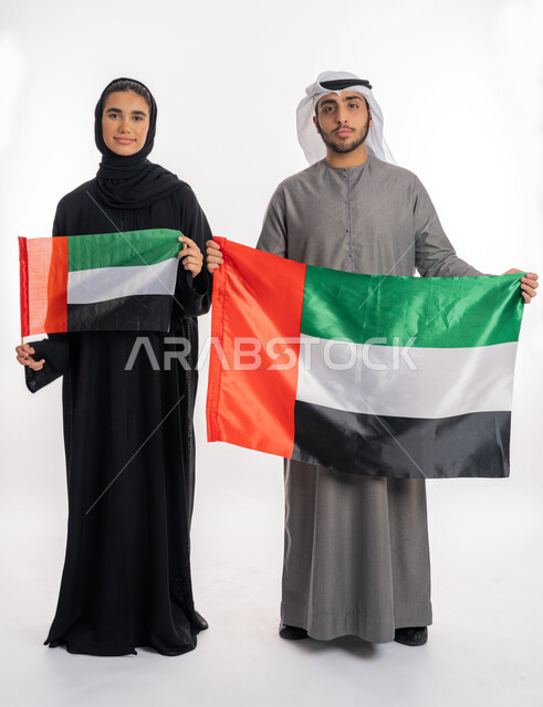 الاحتفال باليوم الوطني الإماراتي٢ ديسمبر، بورتريه لشاب وفتاة عربيان خليجيان إماراتيان مبتسمان يرتديان الزي التقليدي ويحمل كل منهما علم الإمارات بحجمين مختلفين، ذكرى يوم العلم الإماراتي ٣ نوفمبر، الشعور بالفخر والانتماء للوطن، خلفية بيضاء، الطول الكامل للجسم