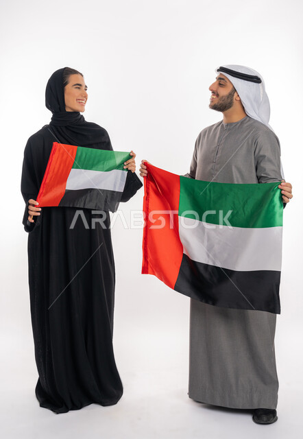 ذكرى اليوم الوطني الإماراتي٢ ديسمبر، بورتريه لشاب وفتاة عربيان خليجيان إماراتيان مبتسمان يرتديان الزي التقليدي ينظران إلى بعضهما ويحملان بيديهما علم الإمارات، الاحتفال بيوم العلم الإماراتي٣ نوفمبر، الشعور بالفخر والانتماء للوطن، خلفية بيضاء، الطول الكامل للجسم