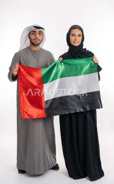 الاحتفال باليوم الوطني الإماراتي٢ ديسمبر، بورتريه لشاب وفتاة عربيان خليجيان إماراتيان مبتسمان يرتديان الزي التقليدي ويحملان بيديهما علم الإمارات، ذكرى يوم العلم الإماراتي ٣ نوفمبر، الشعور بالفرح الافتخار بالوطن، الأعياد والمناسبات الوطنية، خلفية بيضاء، الطول الكامل للجسم
