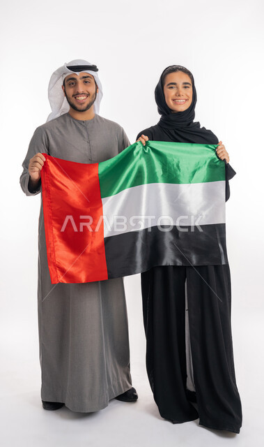الاحتفال باليوم الوطني الإماراتي٢ ديسمبر، بورتريه لشاب وفتاة عربيان خليجيان إماراتيان مبتسمان يرتديان الزي التقليدي ويحملان بيديهما علم الإمارات، ذكرى يوم العلم الإماراتي ٣ نوفمبر، الشعور بالفرح والبهجة، الأعياد والمناسبات الوطنية، خلفية بيضاء، الطول الكامل للجسم
