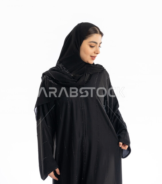 Concern about appearance, concept of femininity and softness, displaying and marketing clothes, Saudi girl looking at something with gestures of joy and pleasure, expressions of self-confidence, portrait of a young Arab Gulf Emirati woman wearing an abaya and hijab, white background