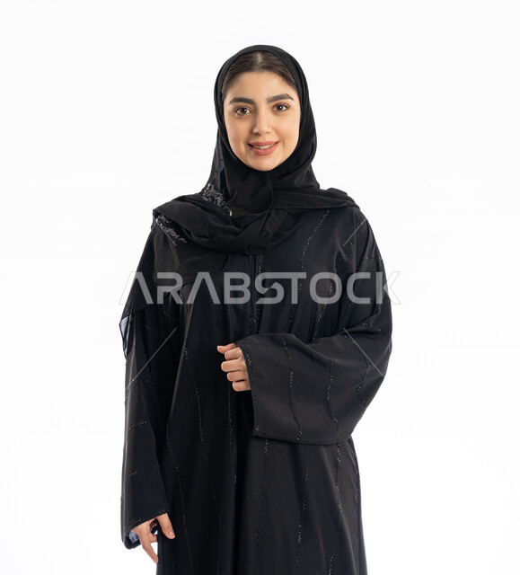 عرض الملابس والتسويق لها، الاهتمام بالمظهر الخارجي، فتاة سعودية تنظر إلى الكاميرا بإيماءات البهجة والسرور، مفهوم الأنوثة والنعومة، تعابير الثقة بالنفس، بورتريه لشابة عربية خليجية إماراتيه ترتدي العباية والحجاب، خلفية بيضاء
