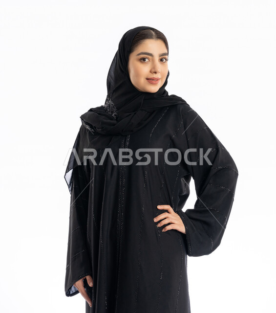 الاعتناء بالمظهر الخارجي، عارضة أزياء سعودية تنظر إلى الكاميرا بإيماءات البهجة والسرور، مفهوم الأنوثة والنعومة، تعابير الثقة بالنفس، بورتريه لشابة عربية خليجية إماراتيه ترتدي العباية والحجاب تقوم بوضع يدها على خصرها، خلفية بيضاء