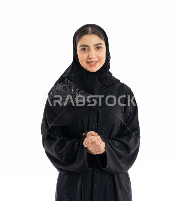 مفهوم الأنوثة والنعومة، تعابير التردد في طلب شيء ما، الاهتمام بالمظهر الخارجي، بورتريه لشابة عربية خليجية إماراتيه ترتدي العباية والحجاب تقوم بشبك يديها ببعضها، فتاة سعودية أنيقة تنظر إلى الكاميرا بإيماءات البهجة والسرور، خلفية بيضاء