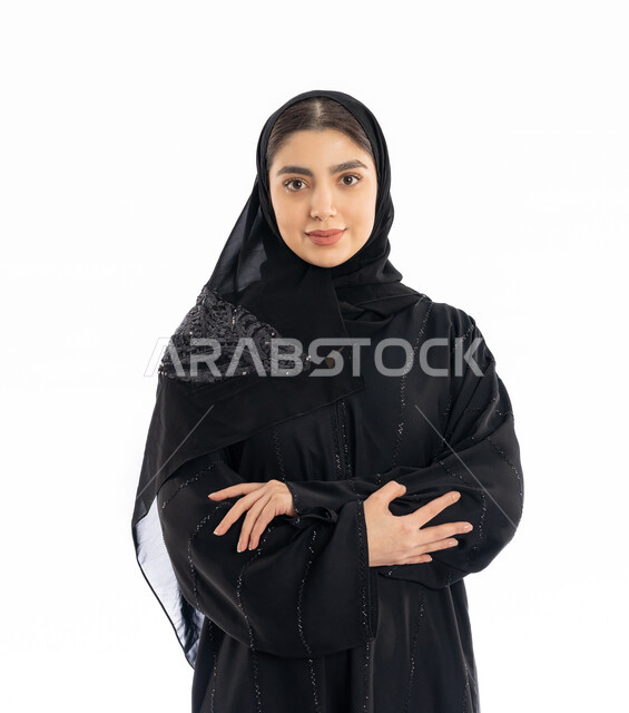 Concern about appearance, concept of femininity and softness, expressions of self-confidence, close-up portrait of a young Arab Gulf Emirati woman wearing an abaya and hijab standing with folded hands, elegant Saudi girl looking at the camera with gestures of joy and pleasure, white background