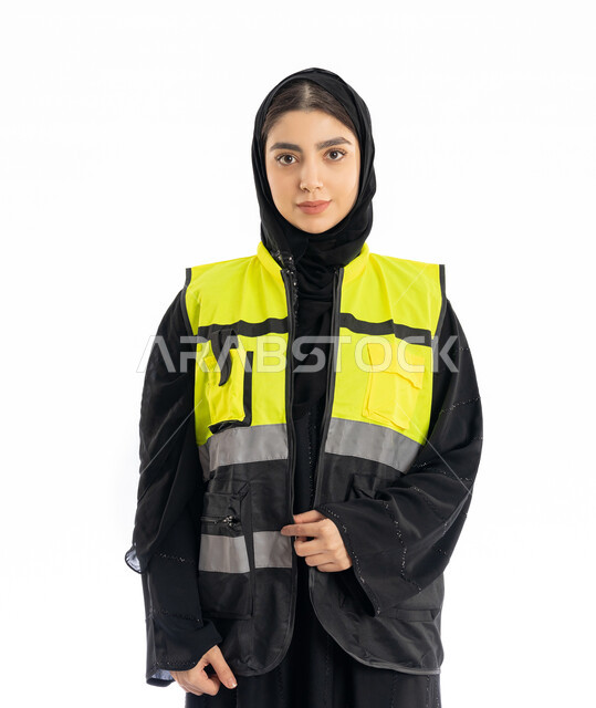 متابعة سير المشاريع، مهن ووظائف هندسية في الامارات، مشاركة النساء في العمل، بورتريه لمهندسة عربية خليجية إماراتية ترتدي العباية والحجاب وسترة الحماية، شابة سعودية تقف باستقامة وتنظر إلى الكامير بإيماءات الحزم والجدية، خلفية بيضاء