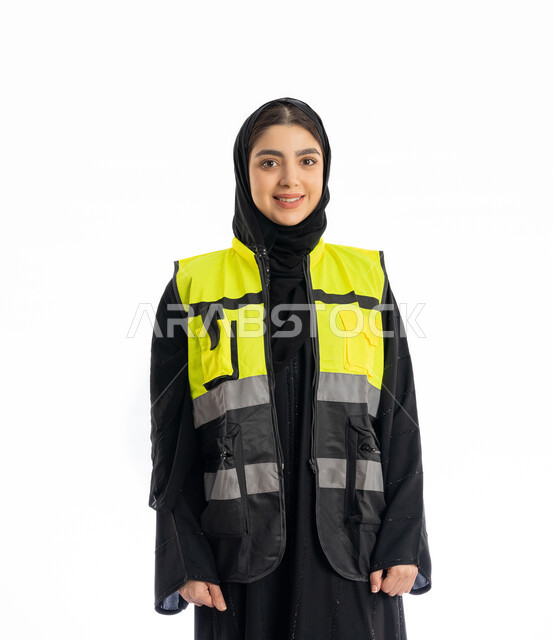 مهن ووظائف هندسية في الامارات، مشاركة النساء في العمل، متابعة سير المشاريع، بورتريه لمهندسة عربية خليجية إماراتية ترتدي العباية والحجاب وسترة الحماية، شابة سعودية تقف باستقامة وتنظر إلى الكامير بإيماءات البهجة والسرور، خلفية بيضاء