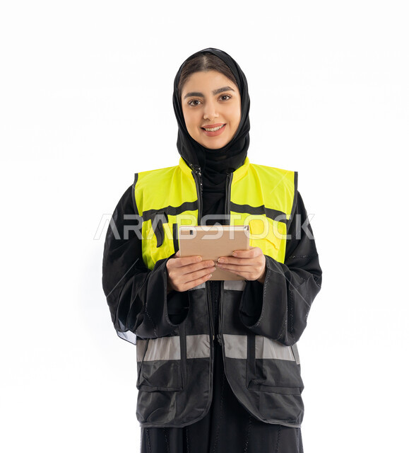 مهن ووظائف هندسية نسائية في الامارات، النظر إلى الكامير بإيماءات البهجة والسرور، تدوين وتسجيل الملاحظات، بورتريه لمهندسة عربية خليجية إماراتية ترتدي العباية والحجاب وسترة الحماية تمسك بيدها دفتر صغير، شابة سعودية تقوم بمتابعة سير المشاريع، خلفية بيضاء