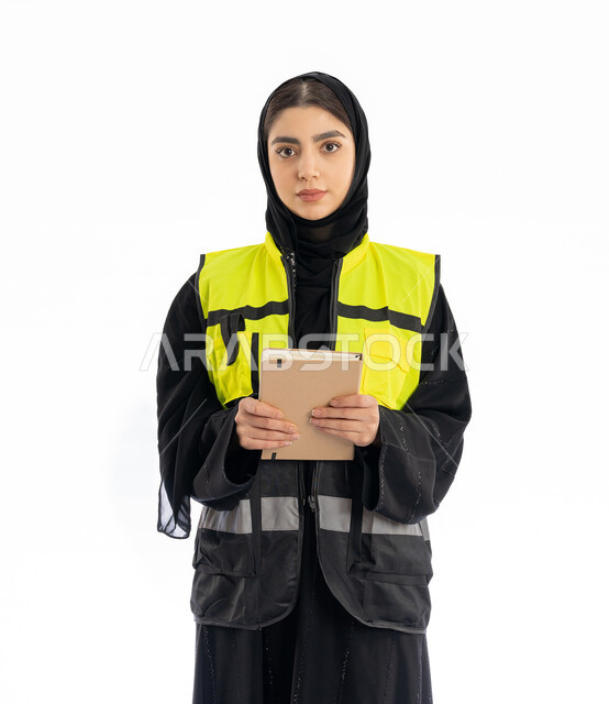 Taking notes, portrait of an Arab Gulf Emirati female engineer wearing an abaya, hijab and a protective vest, holding a small notebook, looking at the camera with gestures of self-confidence, female engineering professions and jobs in the Emirates, a young Saudi woman following up on the progress of projects, white background