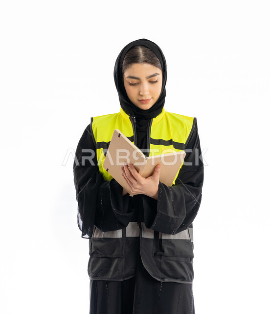 ايماءات التركيز في كتابة شيء ما، متابعة سير المشاريع، مهن ووظائف هندسية في الامارات، مشاركة النساء في العمل، بورتريه لمهندسة عربية خليجية إماراتية ترتدي العباية والحجاب وسترة الحماية، شابة سعودية تقوم بتدوين ونسجيل الملاحظات، خلفية بيضاء