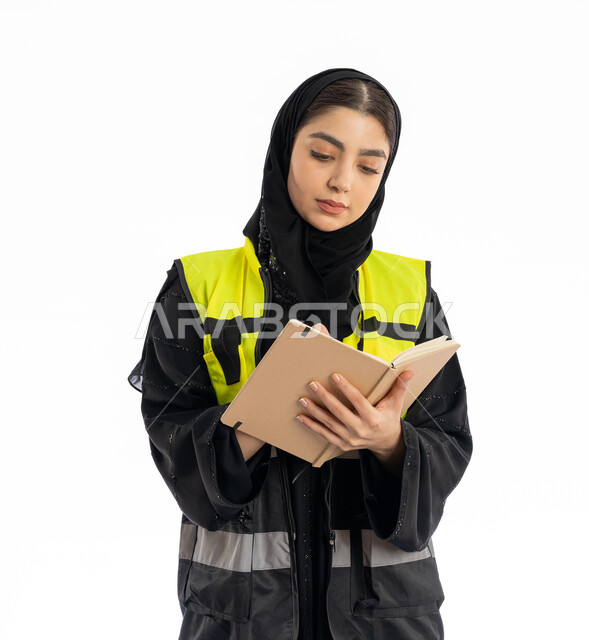 متابعة سير المشاريع، مهن ووظائف هندسية في الامارات، ايماءات التركيز في كتابة شيء ما، مشاركة النساء في العمل، بورتريه مقرب لمهندسة عربية خليجية إماراتية ترتدي العباية والحجاب وسترة الحماية، شابة سعودية تقوم بتدوين ونسجيل الملاحظات، خلفية بيضاء