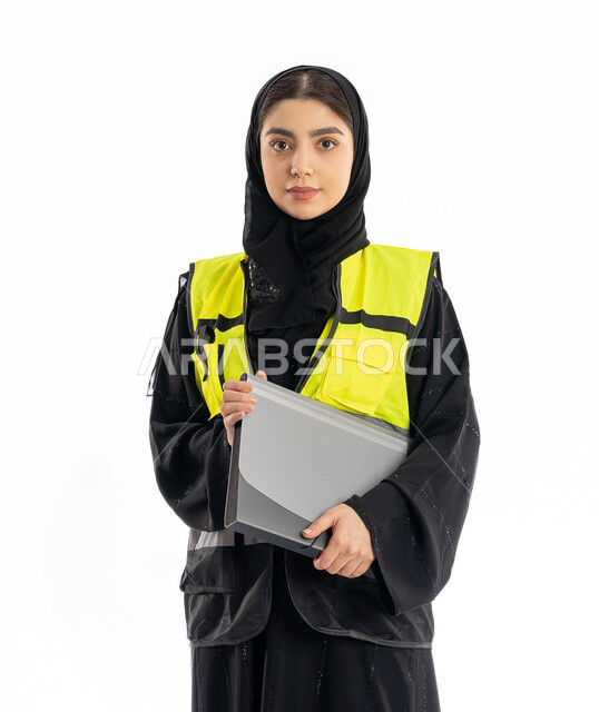 النظر إلى الكاميرا بإيماءات الحزم والجدية، متابعة سير المشاريع، مهن ووظائف هندسية في الامارات، مشاركة النساء في العمل، بورتريه لمهندسة عربية خليجية إماراتية ترتدي العباية والحجاب وسترة الحماية، شابة سعودية تمسك بملف المشروع، خلفية بيضاء