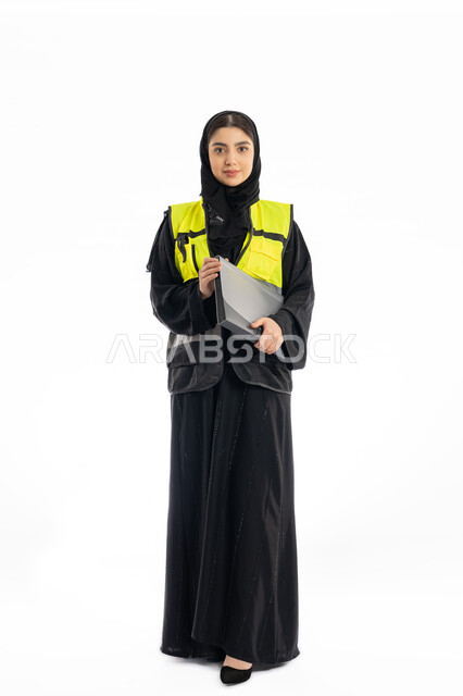 مهن ووظائف هندسية في الامارات، متابعة سير المشاريع، النظر إلى الكاميرا بإيماءات الحزم والجدية، مشاركة النساء في العمل، بورتريه لمهندسة عربية خليجية إماراتية ترتدي العباية والحجاب وسترة الحماية، شابة سعودية تمسك بملف المشروع، صورة لكامل الجسم، خلفية بيضاء