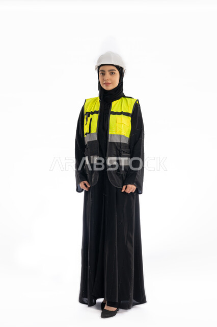 مهن ووظائف هندسيه، مفهوم الهندسه المعماريه, متابعة سير المشاريع والإشراف عليها، بورتريه لمهندسة عربية خليجية اماراتية ترتدي العباية وسترة وخوذة الحماية, فتاة سعودية محجبة تقف باسقامة وتنظر إلى الكاميرا بإيماءات الحزم والجدية, صورة لكامل الجسم، خلفية بيضاء