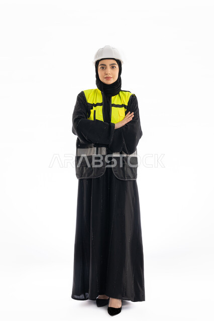 مهن ووظائف هندسية في الامارات، مشاركة النساء في العمل، بورتريه لمهندسة عربية خليجية إماراتية ترتدي العباية والحجاب وسترة وخوذة الحماية، شابة سعودية تقف بأيدي مكتوفة تنظر إلى الكاميرا بإيماءات الحزم والجدية، صورة لكامل الجسم، خلفية بيضاء