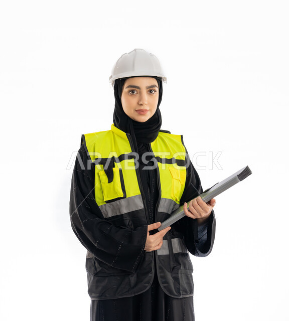 وظائف هندسية في الامارات، النظر إلى شيء ما بإيماءات الحزم والجدية، مشاركة النساء في العمل، متابعة سير المشاريع، بورتريه لمهندسة عربية خليجية إماراتية ترتدي العباية والحجاب وسترة وخوذة الحماية، شابة سعودية تمسك ملف المشروع، خلفية بيضاء
