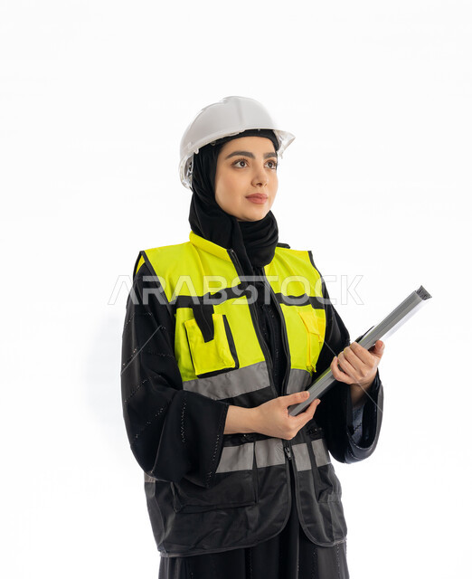 Looking at something with gestures of joy and pleasure, engineering professions and jobs in the Emirates, women's participation in work, following up on the progress of projects, portrait of an Arab Gulf Emirati female engineer wearing an abaya, hijab, jacket and protective helmet, a young Saudi woman holding a project file, white background