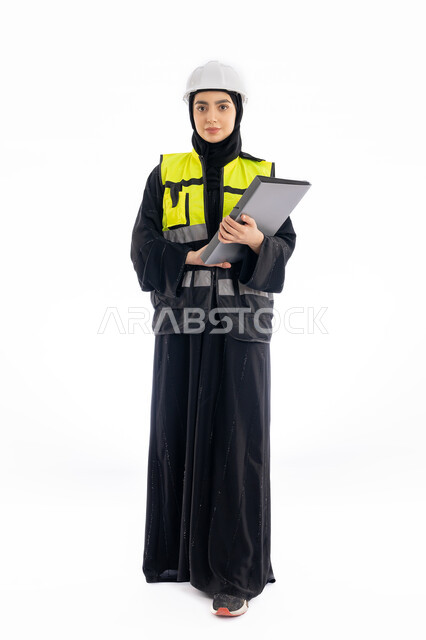 متابعة سير المشاريع، وظائف هندسية في الامارات، النظر إلى الكاميرا بإيماءات الحزم والجدية، مشاركة النساء في العمل، بورتريه لمهندسة عربية خليجية إماراتية ترتدي العباية والحجاب وسترة وخوذة الحماية، شابة سعودية تمسك بملف المشروع، صورة لكامل الجسم، خلفية بيضاء