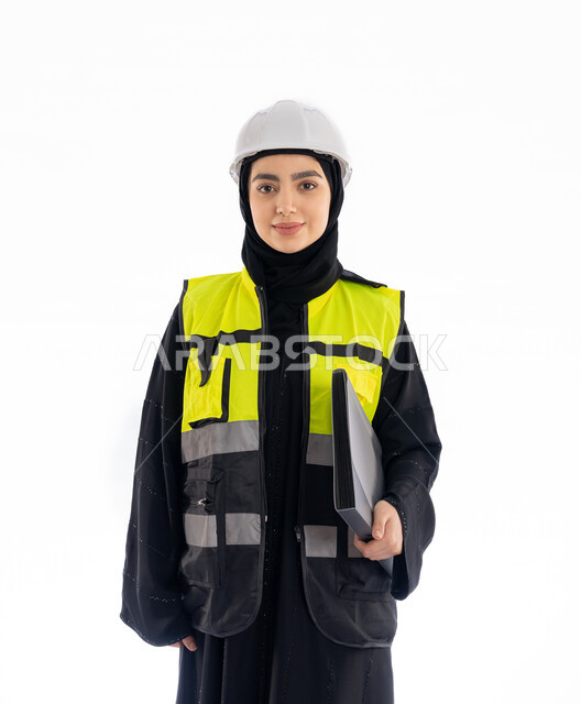مشاركة النساء في العمل، مهن ووظائف هندسية في الامارات، متابعة سير المشاريع، النظر إلى الكاميرا بإيماءات البهجة والسرور، بورتريه لمهندسة عربية خليجية إماراتية ترتدي العباية والحجاب وسترة وخوذة الحماية، شابة سعودية تمسك ملف المشروع، خلفية بيضاء