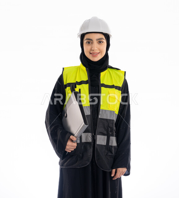 مهن ووظائف هندسية في الامارات، متابعة سير المشاريع، النظر إلى الكاميرا بإيماءات البهجة والسرور، مشاركة النساء في العمل، بورتريه لمهندسة عربية خليجية إماراتية ترتدي العباية والحجاب وسترة وخوذة الحماية، شابة سعودية تمسك ملف المشروع، خلفية بيضاء