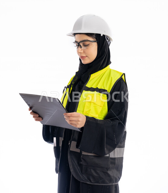 مهن ووظائف هندسية في الامارات، مشاركة النساء في العمل، متابعة سير المشاريع، بورتريه لمهندسة عربية خليجية إماراتية ترتدي العباية والحجاب وسترة الحماية، شابة سعودية تقف باستقامة وتنظر إلى الكامير بإيماءات البهجة والسرور، خلفية بيضاء