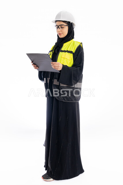 مهن ووظائف هندسية في الامارات، مشاركة النساء في العمل، متابعة سير المشاريع، بورتريه لمهندسة عربية خليجية إماراتية ترتدي العباية والحجاب وسترة الحماية، شابة سعودية تقف باستقامة وتنظر إلى الكامير بإيماءات البهجة والسرور، خلفية بيضاء