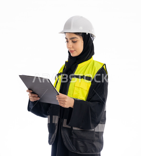 مهن ووظائف هندسية في الامارات، مشاركة النساء في العمل، متابعة سير المشاريع، بورتريه لمهندسة عربية خليجية إماراتية ترتدي العباية والحجاب وسترة الحماية، شابة سعودية تقف باستقامة وتنظر إلى الكامير بإيماءات البهجة والسرور، خلفية بيضاء