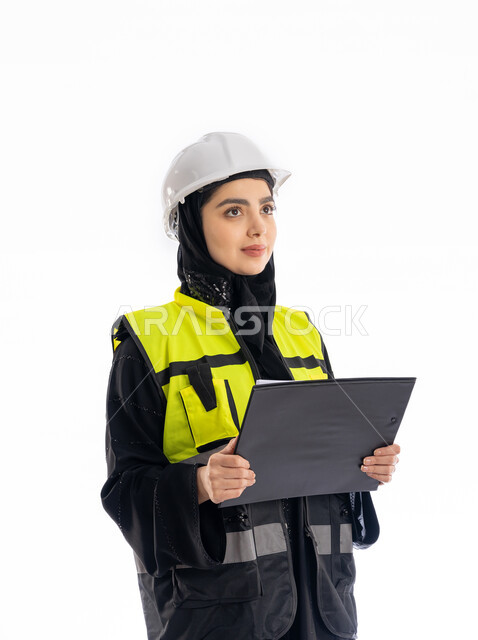 مشاركة النساء في العمل، مهن ووظائف هندسية في الامارات، متابعة سير المشاريع، بورتريه لمهندسة عربية خليجية إماراتية ترتدي العباية والحجاب وسترة ونظارات الحماية تمسك بأوراق الملاحظات الملاحظات، شابة سعودية تنظر إلى شيء ما بإيماءات التركيز، خلفية بيضاء