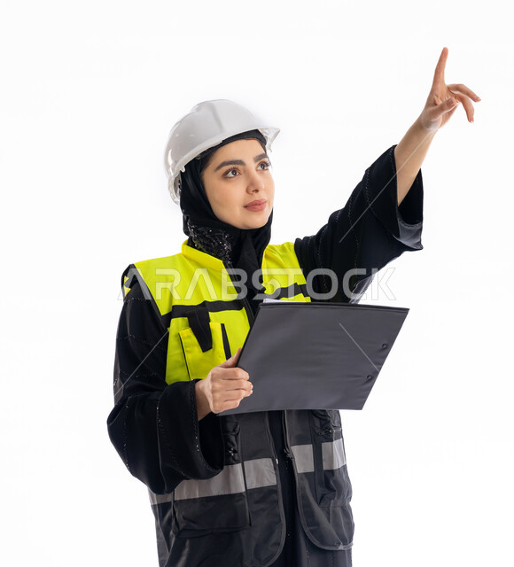 Engineering jobs in the Emirates, women’s participation in the work, monitoring the progress of projects, a portrait of an Arab Gulf Emirati engineer wearing an abaya, hijab, jacket and safety glasses holding a notebook, a young Saudi woman pointing to something with focused gestures, a white background
