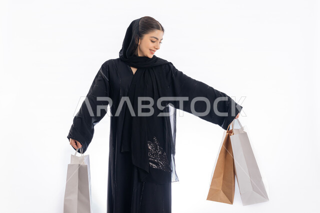 استلام طلبات اونلاين، مفهوم التسوق واقتناء الهدايا، الاستفادة من العروض في شراء الحاجيات الضرورية، فتاة سعودية تنظر إلى الأغراض بإيماءات البهجة والسرور، بورتريه لمرأة عربية خليجية إماراتيه ترتدي العباية والحجاب تمسك بيدها أكياس ورقية، صورة لكامل الجسم، خلفية بيضاء