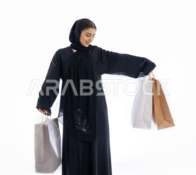 الاستفادة من العروض في شراء الحاجيات الضرورية، استلام طلبات اونلاين، مفهوم التسوق واقتناء الهدايا، فتاة سعودية تنظر إلى الأغراض بإيماءات البهجة والسرور، بورتريه لمرأة عربية خليجية إماراتيه ترتدي العباية والحجاب تمسك بيدها أكياس ورقية، صورة لكامل الجسم، خلفية بيضاء