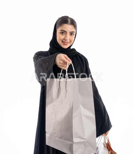 الاستفادة من العروض في شراء الحاجيات الضرورية، مفهوم تقديم الهدايا، استلام طلبات اونلاين، فتاة سعودية تنظر إلى الكاميرا بتعابير البهجة والسرور، بورتريه لمرأة عربية خليجية إماراتيه ترتدي العباية والحجاب تمسك بيدها أكياس ورقية، خلفية بيضاء