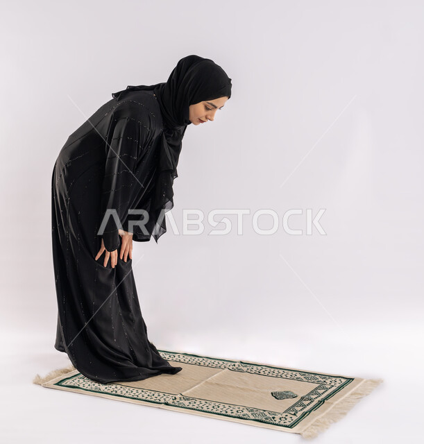 أركان الإسلام الخمسة، مفهوم العبادة والتقرب إلى الله، فتاة سعودية تقوم بأداء فريضة الصلاة، بورتريه لشابة عربية خليجية اماراتية ترتدي العباية والحجاب تقف على سجادة مخصصة تقوم بالركوع، صورة لكامل الجسم، خلفية بيضاء
