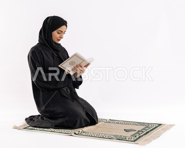 تدبر آيات المصحف الشريف، مفهوم العبادة والتقرب إلى الله، بورتريه لشابة عربية خليجية سعودية محجبة ترتدي العباية تجلس على سجادة مخصصة، فتاة سعودية تقوم بتلاوة القرآن الكريم، صورة لكامل الجسم، خلفية بيضاء