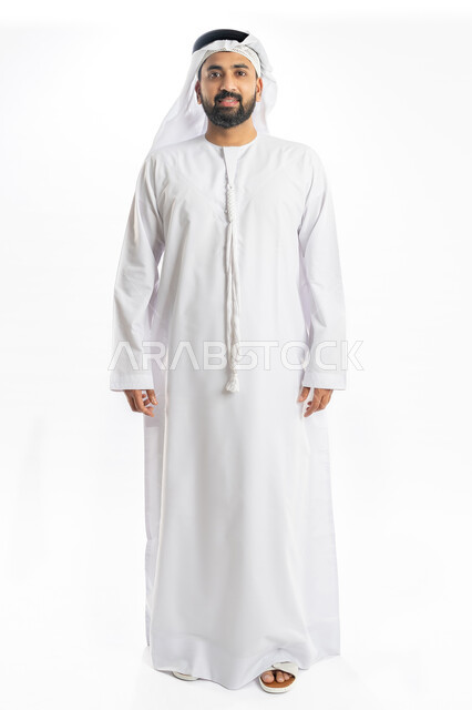 مفهوم الرجولة والقوة، الاهتمام بالمظهر الخارجي، بورتريه لرجل عربي خليجي اماراتي يرتدي الكندورة الاماراتية والغترة يقف باستقامة وينظر الى الكاميرا بإيماءات البهجة والسرور، صورة لكامل الجسم، خلفية بيضاء