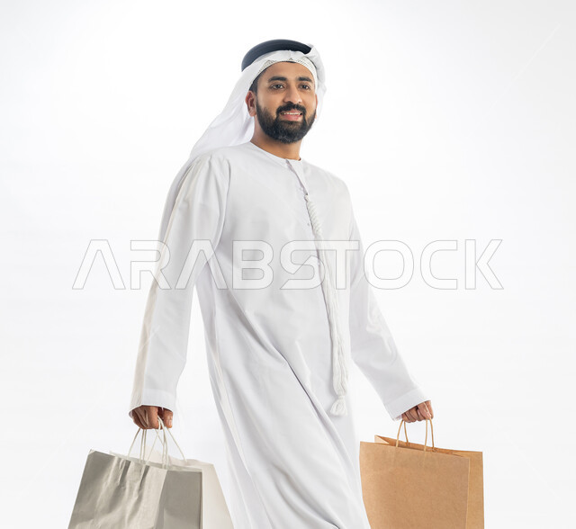 استلام طلبات اونلاين، الاستمتماع في التسوق وشراء الحاجيات والهدايا، النظر إلى الكاميرا بتعابير البهجة والسرور، الاستفادة من العروض في شراء الحاجيات الضرورية، بورتريه من الجانب لرجل عربي خليجي اماراتي يرتدي الكندورة والغترة يمسك بيده أكياس ورقية، خلفية بيضاء