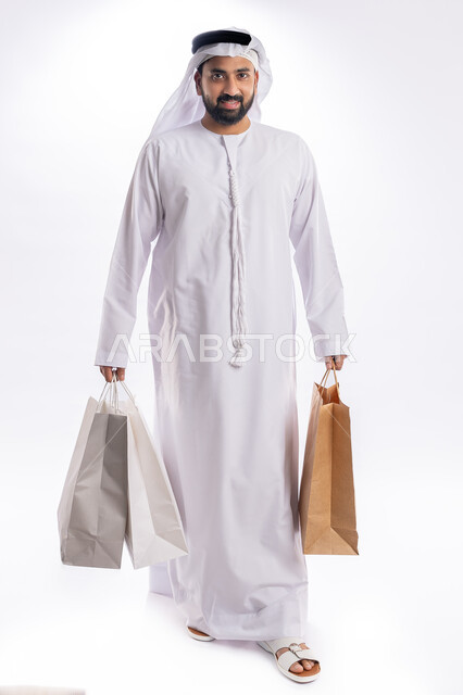الاستفادة من العروض في شراء الحاجيات الضرورية، اقتناء الهدايا والتسوق، بورتريه رجل عربي خليجي اماراتي يرتدي الكندورة والغترة يمسك بيده أكياس ورقية، استلام طلبات اونلاين، النظر إلى الكاميرا بتعابير البهجة والسرور، صورة لكامل الجسم، خلفية بيضاء