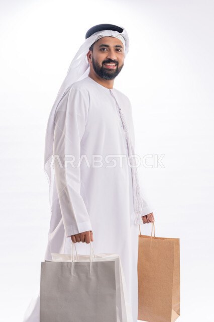 Looking at the camera with joy and pleasure expressions, benefiting from offers in buying necessary needs, acquiring gifts and shopping, portrait from the side to an Emirati Gulf Arab man wearing the kandora and ghutra holds with his hand paper bags, receiving online requests, white background