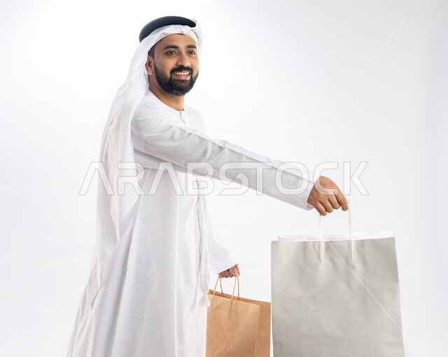 مفهوم تقديم الهدايا، النظر إلى الكاميرا بتعابير البهجة والسرور، الاستفادة من العروض في شراء الحاجيات الضرورية، بورتريه من الجانب لرجل عربي خليجي اماراتي يرتدي الكندورة والغترة يمسك بيده أكياس ورقية، استلام طلبات اونلاين، خلفية بيضاء