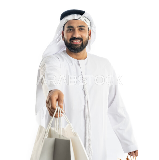 مفهوم تقديم الهدايا، استلام طلبات اونلاين، النظر إلى الكاميرا بتعابير البهجة والسرور، الاستفادة من العروض في شراء الحاجيات الضرورية، بورتريه لرجل عربي خليجي اماراتي يرتدي الكندورة والغترة يمسك بيده أكياس ورقية، خلفية بيضاء