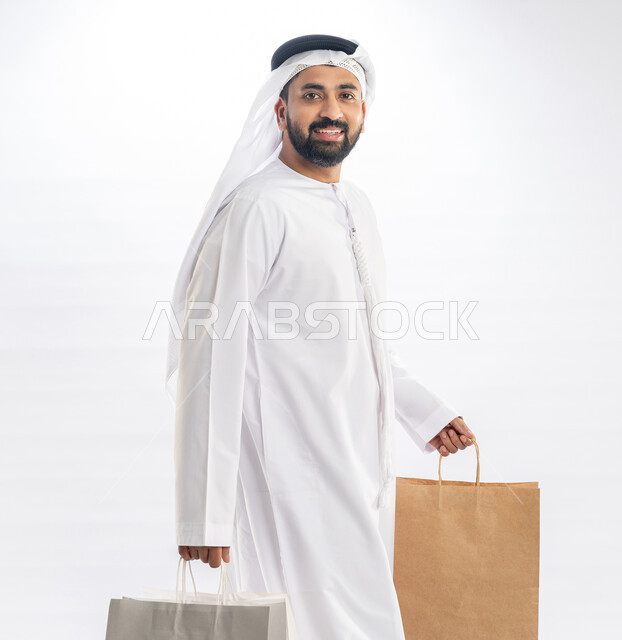 التسوق وشراء الحاجيات والهدايا، الاستفادة من العروضات والتخفيضات، النظر إلى الكاميرا بإيماءات البهجة والسرور، بورتريه لرجل عربي خليجي إماراتي مبتسم يرتدي الكندورة  والغترة يحمل بيديه أكياس ورقية، خلفية بيضاء