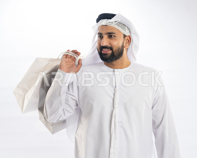 Taking advantage of offers and discounts, looking at something with gestures of joy and pleasure, enjoying shopping and buying necessities and gifts, close-up portrait of a smiling Emirati Gulf Arab man wearing a kandura and ghutra holding paper bags in his hand, white background