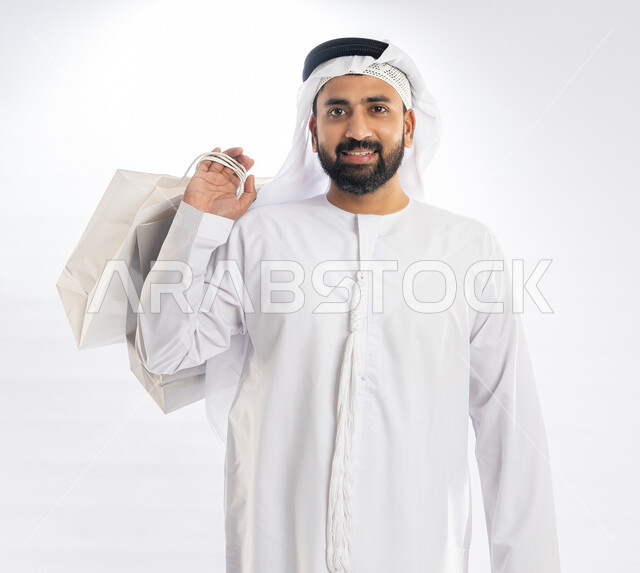 النظر إلى الكاميرا بإيماءات البهجة والسرور، الاستمتاع في التسوق وشراء الحاجيات والهدايا، الاستفادة من العروضات والتخفيضات، بورتريه لرجل عربي خليجي إماراتي مبتسم يرتدي الكندورة  والغترة يحمل بيده أكياس ورقية، خلفية بيضاء