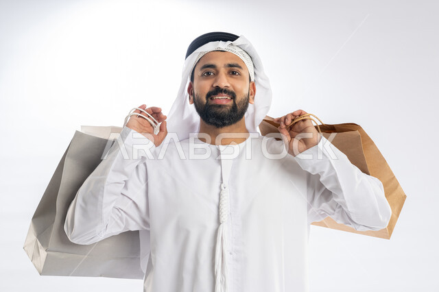 استلام طلبات الاونلاين، الاستفادة من العروض في شراء الحاجيات الضرورية، متعة التسوق واقتناء المشتريات، بورتريه مقرب لرجل عربي خليجي إماراتي يرتدي الكندورة والغترة يمسك بيديه أكياس ورقية يقوم بالنظر إلى الكاميرا بإيماءات البهجة والسرور، خلفية بيضاء