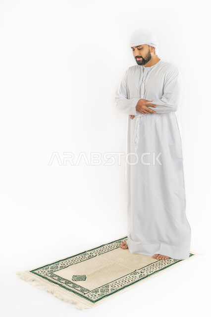 العبادة والتقرب إلى الله، أركان الإسلام الخمسة، الخشوع أثناء أداء فريضة الصلاة، بورتريه لرجل عربي خليجي إماراتي مسلم يرتدي الكندورة والعمامة يقف على سجادة مخصصة، صورة لكامل الجسم، خلفية بيضاء