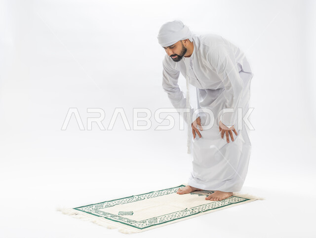 أركان الإسلام الخمسة، مفهوم العبادة والتقرب إلى الله، الخشوع أثناء أداء فريضة الصلاة، بورتريه لرجل عربي خليجي إماراتي مسلم يرتدي الكندورة والعمامة يقف على سجادة مخصصة يقوم بالركوع، صورة لكامل الجسم، خلفية بيضاء
